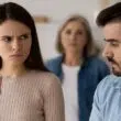 Une femme en couple lève la main pour stopper une discussion tendue, avec la présence d’une mère en arrière-plan, illustrant l’impact d’une mère toxique sur une relation amoureuse.