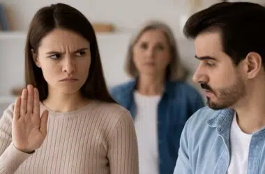Une femme en couple lève la main pour stopper une discussion tendue, avec la présence d’une mère en arrière-plan, illustrant l’impact d’une mère toxique sur une relation amoureuse.